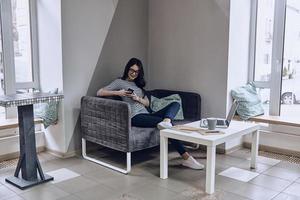 sms'en naar bedrijf partner. mooi jong glimlachen vrouw in eyewear gebruik makend van slim telefoon terwijl zittend Aan de sofa Bij haar werken plaats foto