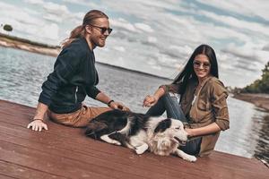 uitgeven tijd samen. mooi jong paar spelen met hond terwijl zittend in de buurt de meer buitenshuis foto