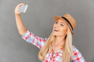 funky selfie. vrolijk jong vrouw maken selfie Aan haar slim telefoon terwijl staand tegen grijs achtergrond foto