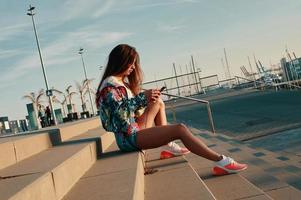 sms'en naar vriend. aantrekkelijk jong vrouw in sport- kleding gebruik makend van slim telefoon terwijl zittend Aan de stappen buitenshuis foto