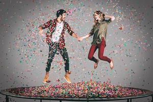 zorgeloos plezier. in de lucht schot van mooi jong vrolijk paar Holding handen terwijl jumping Aan trampoline samen met confetti allemaal in de omgeving van hen foto