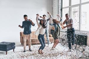 gaan gek. vol lengte van gelukkig jong mensen dansen terwijl uitgeven tijd Bij huis met confetti vliegend overal foto