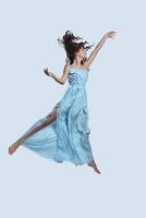 bereiken de lucht. vol lengte studio schot van aantrekkelijk jong vrouw in elegant jurk gebaren terwijl zweven tegen grijs achtergrond foto
