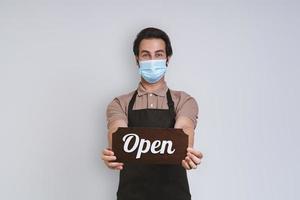 knap jong Mens in schort vervelend beschermend gezicht masker en Holding Open teken terwijl staand tegen grijs achtergrond foto