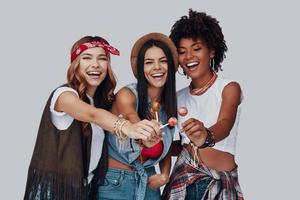 drie aantrekkelijk jong Dames aan het eten lolly en glimlachen terwijl staand tegen grijs achtergrond foto