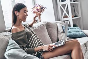 modern technologieën maken haar gelukkig. aantrekkelijk jong Aziatisch vrouw gebruik makend van laptop en glimlachen terwijl zittend Aan de sofa Bij huis foto