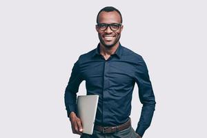 zelfverzekerd zakenman. knap jong Afrikaanse Mens draag- laptop en op zoek Bij camera met glimlach terwijl staand tegen grijs achtergrond foto