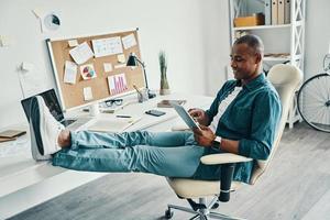 freelance werk. knap jong Afrikaanse Mens in overhemd gebruik makend van digitaal tablet en glimlachen terwijl zittend in de kantoor foto