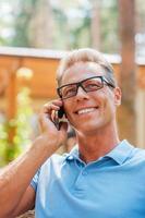 sharing mooi zo nieuws met vrienden. gelukkig volwassen Mens pratend Aan de mobiel telefoon en glimlachen terwijl zittend buitenshuis met huis in de achtergrond foto