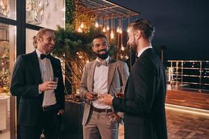 drie goed gekleed mannen drinken whisky en communiceren terwijl uitgeven tijd Aan partij foto