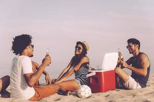 ontspannende met het beste vrienden. vrolijk jong mensen uitgeven mooi hoor tijd samen terwijl zittend Aan de strand en drinken bier foto