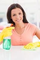 huisvrouw Bij werk. jong mooi vrouw in geel handschoenen Holding een vod en verstuiven fles terwijl glimlachen Bij camera foto