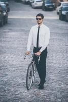 zijn elegant rijden. vol lengte van knap goed gekleed jong Mens in zonnebril rollend zijn fiets buitenshuis foto