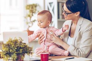 werken samen met vreugde. vrolijk jong mooi zakenvrouw op zoek Bij haar baby meisje met glimlach terwijl zittend Bij haar werken plaats foto