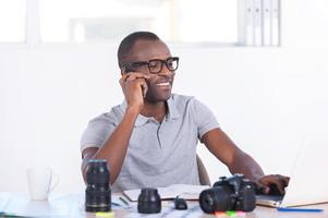 werken Aan creatief project. knap jong Afrikaanse Mens in gewoontjes slijtage zittend Bij zijn werken plaats en pratend Aan de mobiel telefoon foto