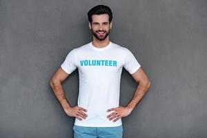 het beste vrijwilliger. zelfverzekerd jong Mens in vrijwilliger t-shirt houden handen Aan heupen en op zoek Bij camera met glimlach terwijl staand tegen grijs achtergrond foto