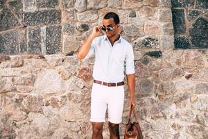 koel en stijlvol. zelfverzekerd jong Afrikaanse Mens in slim gewoontjes kleren draag- zak en op zoek weg terwijl aanpassen zijn zonnebril en staand tegen de stoned muur buitenshuis foto