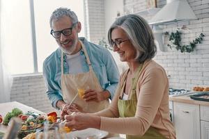 vrolijk senior paar in schorten voorbereidingen treffen gezond avondeten en glimlachen terwijl uitgeven tijd Bij huis foto