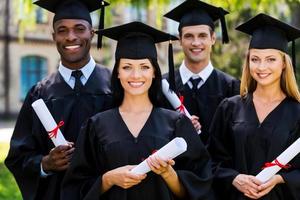 gevoel zelfverzekerd in hun toekomst. vier college afgestudeerden in diploma uitreiking jurken staand dichtbij naar elk andere en glimlachen