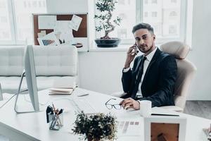 altijd verkrijgbaar. echt jong Mens in formele kleding pratend Aan de telefoon en op zoek weg terwijl zittend Bij de kantoor bureau foto