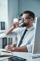 knap jong Mens in overhemd en stropdas drinken water terwijl zittend in de kantoor foto