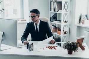 bezig Bij werk. knap jong Mens in vol pak werken met documenten terwijl zittend Bij de kantoor bureau foto