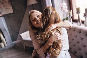 het beste vrienden. top visie van aantrekkelijk jong Dames in elegant jurken omarmen en glimlachen terwijl zittend Aan de sofa foto