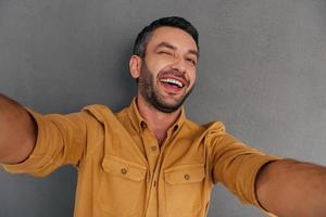 selfie plezier. glimlachen jong Mens maken selfie en grimassen terwijl staand tegen grijs achtergrond foto