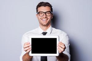 kopiëren ruimte Aan zijn tablet. knap jong Mens in overhemd en stropdas tonen zijn digitaal tablet en glimlachen terwijl staand tegen grijs achtergrond foto