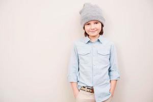 zo jong en zo elegant knap weinig jongen in hoed Holding handen in zakken en op zoek Bij camera terwijl staand tegen grijs achtergrond foto