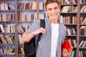 slim en zelfverzekerd leerling. knap jong Mens Holding boeken en glimlachen terwijl staand in bibliotheek foto