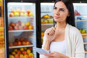 vrouw met boodschappen doen lijst. attent jong vrouw Holding boodschappen doen lijst en op zoek weg terwijl staand in voorkant van koelkasten in kruidenier op te slaan foto