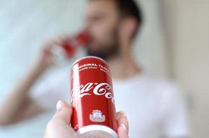 Charkov. Oekraïne - mei 2, 2019 Kaukasisch Mens drankjes Coca Cola drinken in garage interieur en mannetje hand- presenteert een coca cola rood kan in focus foto
