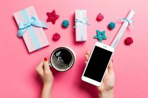 top visie van een vrouw Holding een telefoon in een hand- en een kop van koffie in een ander hand- Aan roze achtergrond. Kerstmis decoraties en speelgoed. nieuw jaar vakantie concept. mockup foto