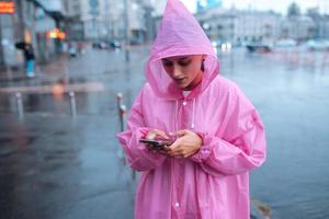 een jong vrouw in een regenjas op zoek Bij de smartphone foto