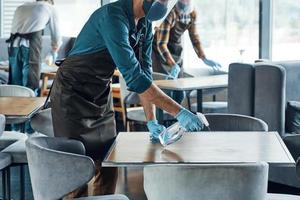 bezig jong mannetje obers in beschermend werkkleding schoonmaak tafels in restaurant foto