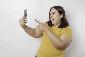 een portret van een gelukkig Aziatisch groot formaat vrouw is glimlachen en Holding haar smartphone vervelend een geel t-shirt geïsoleerd door een wit achtergrond foto