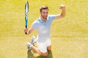 ik won top visie van gelukkig jong Mens in polo overhemd Holding tennis racket en gebaren terwijl geknield Aan tennis rechtbank foto