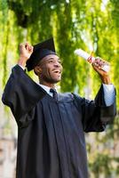 Tenslotte afgestudeerd gelukkig jong Afrikaanse Mens in diploma uitreiking jurken Holding diploma en stijgende lijn armen omhoog