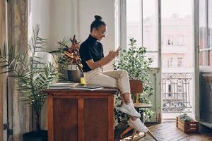 zelfverzekerd jong vrouw gebruik makend van slim telefoon terwijl zittend Aan de houten tafel Bij huis foto