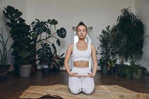 zelfverzekerd jong vrouw in sport- kleding mediteren met kamerplanten allemaal in de omgeving van haar foto