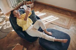 top visie van gelukkig jong vrouw maken selfie door haar slim telefoon terwijl ontspannende in stoel Bij huis foto