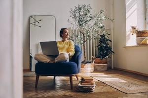 mooi jong vrouw gebruik makend van laptop terwijl zittend in een comfortabel stoel Bij huis foto