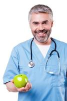 toetreden gezond levensstijl gelukkig volwassen chirurg in blauw uniform Holding groen appel en glimlachen terwijl staand geïsoleerd Aan wit foto