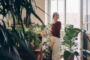 zelfverzekerd jong vrouw gieter kamerplanten en glimlachen terwijl staand Bij de huiselijk kamer foto