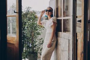 modieus jong vrouw aanpassen haar bril en glimlachen terwijl staand Aan de balkon foto