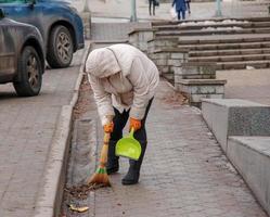 een arbeider met een bezem veegt de straat van stof en brokstukken. de conciërge reinigt de weg. een vrouw schoonmaakster veegt de trottoir met een bezem en stoffer foto