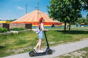 jonge heldere vrouw glimlacht en rijdt elektrische scooter in de zomer in de stad foto