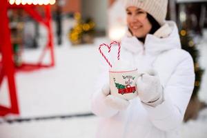vrouw met mok met sneeuw, snoep riet en opschrift vrolijk en helder in haar handen buitenshuis in warm kleren in winter feestelijk markt. fee lichten slingers versierd sneeuw stad- voor nieuw jaar. Kerstmis foto