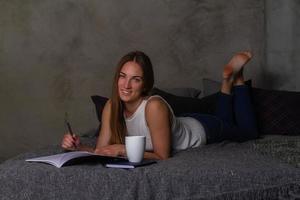 glimlachen vrouw met een notitieboekje en een wit kop aan het liegen Aan haar maag in bed foto